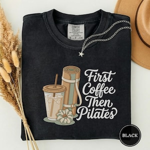 May include: A black crewneck t-shirt featuring a graphic of an iced coffee, a rolled-up yoga mat, and a scrunchie. The text reads First Coffee Then Pilates. The shirt has a Comfort Colors label at the inner neckline and a silver star necklace.