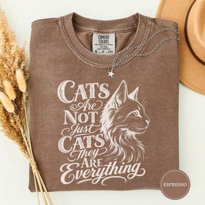 May include: A brown short-sleeved t-shirt with a vintage wash, featuring a white graphic of a cat profile and the text "Cats Are Not Just Cats They Are Everything" printed on the front. The shirt has a crew neckline and a Comfort Colors brand label.