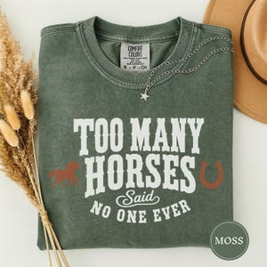 Op de afbeelding: Een mosgroen T-shirt met korte mouwen en een vintage wassing. De voorkant bevat witte tekst met de tekst TOO MANY HORSES Said NO ONE EVER, vergezeld van een klein bruin paard-silhouet en een bruine hoefijzer-afbeelding aan de zijkanten.