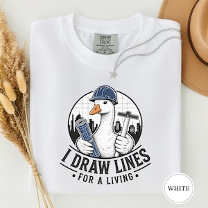 Może przedstawiać: Biały t-shirt z grafiką gęsi w niebieskim kasku budowlanym, trzymającej zwinięty projekt i ekierkę. W tle widać sylwetkę miasta. Napis głosi I DRAW LINES FOR A LIVING czarnymi literami.