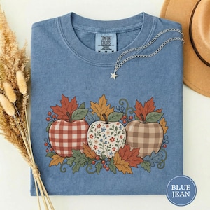 May include: A blue short-sleeve t-shirt featuring a graphic of three apples surrounded by autumn leaves and berries. The apples have red gingham, floral, and tan plaid patterns. The shirt has a Comfort Colors label at the inner neckline.
