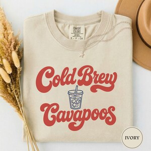 Può includere: Una t-shirt color avorio con la scritta rossa "Cold Brew" sopra un'illustrazione a linee blu di una tazza di caffè, con la scritta rossa "Cavapoos" sotto. La maglietta ha un girocollo e un'etichetta con scritto "Comfort Colors S P CH".