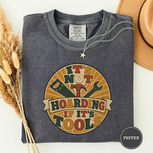 Op de afbeelding: Een antracietgrijs t-shirt met korte mouwen en een ronde afbeelding met een zaag, hamer en moersleutel. De tekst luidt IT'S NOT HOARDING IF IT'S TOOLS. Het shirt heeft een ronde hals, een vintage wassing en een zilveren sterrenketting.