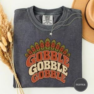 Può includere: Una t-shirt a girocollo grigio antracite con una grafica di una coda di tacchino in verde oliva, arancione e crema. Il testo GOBBLE GOBBLE GOBBLE è stampato in un carattere retrò sotto le piume. La maglietta ha un'etichetta Comfort Colors.