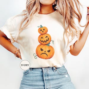 May include: An ivory t-shirt featuring a graphic print of three stacked orange pumpkins with different facial expressions. The top pumpkin has a surprised face, the middle one has a smiling jack-o'-lantern face, and the bottom one has an angry face.