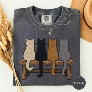 Può includere: Una t-shirt girocollo grigio antracite con la grafica di quattro gatti color crema, nero, marrone e grigio seduti su una panca di legno. La maglietta presenta un'etichetta Comfort Colors sul collo e una collana con ciondolo a stella argento.