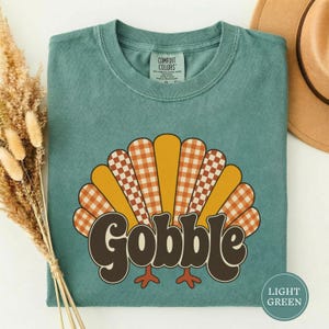 Può includere: Una t-shirt girocollo color verde salvia con la grafica di una coda di tacchino con piume alternate in giallo tinta unita e a quadretti arancioni. La parola GOBBLE è stampata in grandi lettere marrone scuro in stile retrò.