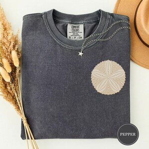 May include: A charcoal gray crew neck t-shirt with a faded wash, featuring a beige sand dollar graphic on the chest. A silver-toned chain necklace with a small star pendant rests on the collar. The inner label reads Comfort Colors.