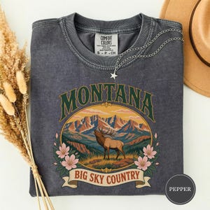 Puede incluir: Una camiseta de cuello redondo gris carbón con un estampado gráfico de un alce marrón en un paisaje montañoso con flores rosas. El texto dice MONTANA BIG SKY COUNTRY. La camiseta tiene un lavado vintage y una etiqueta de la marca Comfort Colors.