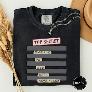 Può includere: Una t-shirt nera a girocollo con una grafica che presenta il testo rosso TOP SECRET sopra cinque barre orizzontali grigie. Il testo Nothing To See Here Move Along è stampato in nero su piccole etichette rettangolari color beige.