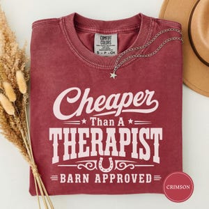 Op de afbeelding: Een karmozijnrood T-shirt met ronde hals, witte tekst Cheaper Than A Therapist Barn Approved en een hoefijzergrafiek. Het shirt heeft een Comfort Colors-label bij de binnenhals en een zilveren sterrenketting die op de stof rust.