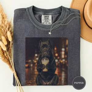 May include: A charcoal grey crew neck t-shirt with a vintage wash finish. The centre features a graphic of a black dog holding a clear plastic cup with a straw. The interior neck label reads COMFORT COLORS. A silver star pendant necklace is included.