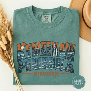 Può includere: Una t-shirt a maniche corte color verde salvia con una stampa grafica che riporta il testo KEWEENAW PENINSULA MICHIGAN. Il design include un paesaggio stilizzato di scogliere, alberi e acqua in tonalità arancione, blu scuro e bianco, con un sole rotondo.