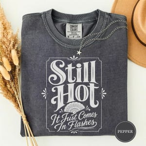 Op de afbeelding: Een antracietgrijs T-shirt met korte mouwen en een vintage wassing. De voorkant heeft witte tekst met de tekst Still Hot It Just Comes In Flashes, vergezeld van een afbeelding van een waaier. Een zilveren ster-halsketting rust op de kraag.