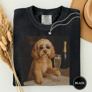 May include: A black crew neck t-shirt featuring a graphic print of a golden-coloured dog wearing oval sunglasses, holding a champagne flute, with a bottle in the background. The shirt has a Comfort Colors label at the inner neckline.