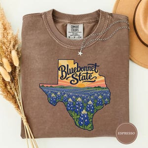May include: A brown short-sleeve t-shirt featuring a graphic of the state of Texas filled with a field of bluebonnet flowers under a sunset sky. The text "Bluebonnet State" is printed in dark blue script over the sunset graphic.