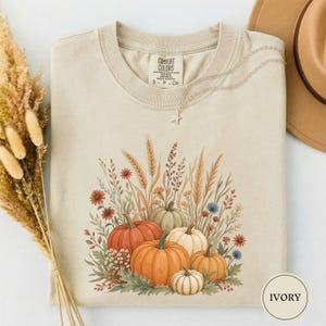 Puede incluir: Una camiseta color marfil con un estampado gráfico de calabazas naranjas, verdes y blancas rodeadas de tallos de trigo y flores silvestres. La etiqueta interior dice Comfort Colors S P CH. Un collar de estrella plateado descansa en el cuello.