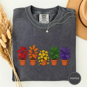 Peut inclure: Un t-shirt à col rond gris anthracite avec une étiquette Comfort Colors au niveau du col. Le t-shirt présente un motif de cinq plantes en pot alignées, colorées en rouge, orange, jaune, vert et violet. Un collier pendentif étoile argenté est posé dessus.