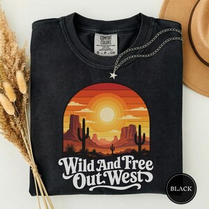 Puede incluir: Una camiseta negra de manga corta con un gráfico de un paisaje desértico con cactus, mesetas y una puesta de sol en tonos naranja, amarillo y rojo. El texto "Wild And Free Out West" está impreso en blanco debajo del gráfico.
