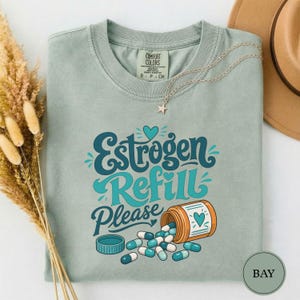 Puede incluir: Una camiseta verde salvia con el texto Estrogen Refill Please en letras azul cerceta y azul claro. El gráfico incluye un frasco de pastillas naranja con un icono de corazón y cápsulas azules y blancas. Se ve un collar de estrella plateado.