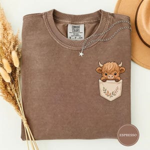 Puede incluir: Una camiseta marrón con lavado mineral que presenta un gráfico de una vaca highland de dibujos animados asomándose por un bolsillo. El diseño del bolsillo incluye una pequeña ilustración de hojas. La camiseta tiene una etiqueta Comfort Colors.