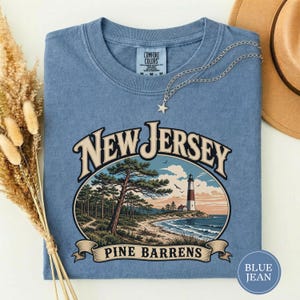 May include: A blue short-sleeve t-shirt featuring a graphic of a lighthouse on a coastline with pine trees and the text New Jersey Pine Barrens. The shirt has a crew neckline and a Comfort Colors label on the inside of the collar area.