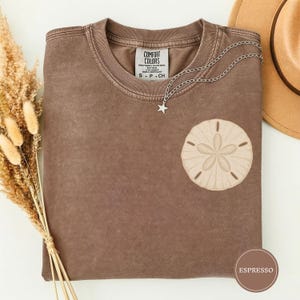 Puede incluir: Una camiseta de manga corta marrón con un lavado desteñido, que presenta un gráfico de dólar de arena beige en el pecho. La camiseta tiene un cuello redondo con una etiqueta Comfort Colors y se complementa con un collar de cadena plateado.
