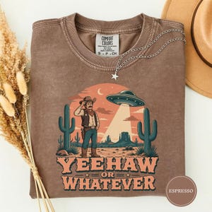 Może przedstawiać: Brązowy t-shirt z okrągłym dekoltem i grafiką przedstawiającą kowboja na pustyni z kaktusami oraz latającym spodkiem powyżej. Napis YEEHAW OR WHATEVER jest wydrukowany dużymi literami pod ilustracją. Koszulka posiada metkę Comfort Colors.
