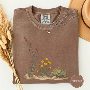 Könnte beinhalten: Ein braunes T-Shirt mit Rundhalsausschnitt und einer Grafik von Wüstenpflanzen, darunter hohe Ocotillo-Stängel mit roten Blüten und Büschel gelber Wildblumen. Das Shirt hat ein Comfort Colors-Etikett am Hals und eine silberne Sternkette.
