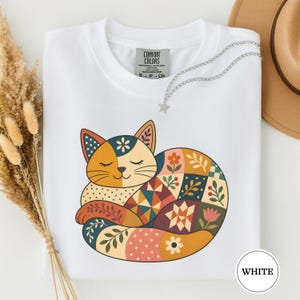 Può includere: Una t-shirt bianca con la grafica di un gatto raggomitolato dal design patchwork. L'illustrazione del gatto incorpora vari motivi, tra cui fiori, foglie, pois e forme geometriche nelle tonalità dell'arancione, del blu e del rosa.