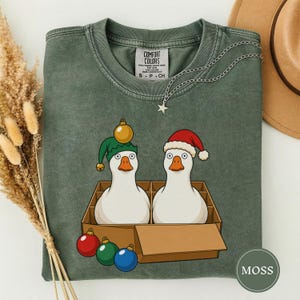 May include: A moss green crewneck t-shirt featuring a graphic of two white ducks in a cardboard box. One duck wears a green elf hat with a gold ornament, the other a red Santa hat. Three colorful ornaments lie in front of the box. The shirt has a Comfort Colors label.