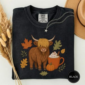 May include: A black crewneck t-shirt featuring a graphic of a brown Highland cow, a pumpkin, and a mug of coffee with whipped cream and a cinnamon stick, surrounded by autumn leaves. The inner label reads Comfort Colors.