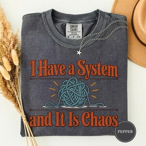 May include: A charcoal gray short-sleeve t-shirt with a graphic print. The text reads I Have a System and It Is Chaos in orange letters, centered above and below a blue yarn ball illustration. The shirt has a crew neck and a tag label.