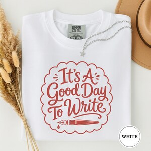 May include: A white short-sleeved t-shirt with a red graphic print featuring a scalloped circular border, a fountain pen illustration, and the text "It's A Good Day To Write". The shirt has a Comfort Colors brand label at the inner neckline.