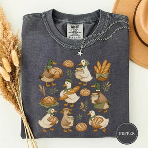 Może przedstawiać: Ciemnoszary t-shirt z okrągłym dekoltem i nadrukiem przedstawiającym różne kaczki trzymające bochenki chleba, bagietki oraz szklane słoiki. Koszulka posiada metkę Comfort Colors przy wewnętrznym dekolcie oraz naszyjnik z wisiorkiem w kształcie gwiazdy.