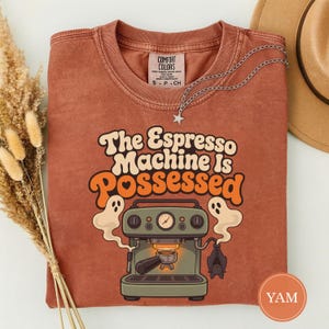 May include: A rust-colored t-shirt featuring a graphic of an espresso machine with two ghosts and a bat. The text reads The Espresso Machine Is Possessed. The shirt has a crew neckline and a Comfort Colors brand label on the inside collar.