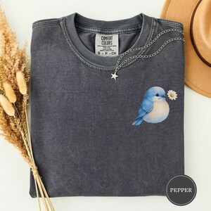 May include: A charcoal grey short-sleeve t-shirt with a crew neckline and a Comfort Colors label. The shirt features a graphic of a small blue bird holding a white daisy in its beak. A silver-toned star charm necklace rests on the fabric.