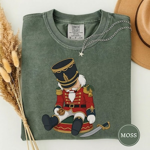 Könnte beinhalten: Ein moosgrünes T-Shirt mit Rundhalsausschnitt und einer Grafik eines Nussknackers in roter Uniform mit goldenen Akzenten, der sitzt und eine Walnuss hält. Eine silberne Sternkette liegt am Ausschnitt. Das Etikett zeigt Comfort Colors S P CH.
