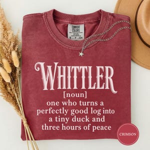 Op de afbeelding: Een karmozijnrood t-shirt met ronde hals en witte tekst: Whittler [noun] one who turns a perfectly good log into a tiny duck and three hours of peace. Het shirt heeft een Comfort Colors label in de nek en een zilverkleurige sterrenketting.