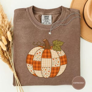 May include: A brown crewneck t-shirt featuring a large graphic of a pumpkin made of patchwork squares in orange, white, and floral patterns. The shirt has a tag that reads Comfort Colors. A silver star necklace rests on the neckline of the shirt.