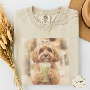 Peut inclure: Un t-shirt ivoire avec une impression graphique représentant un chien brun doré au pelage duveteux, portant un petit nœud sur la tête et tenant un gobelet en plastique transparent rempli d'une boisson verte. Col rond et étiquette Comfort Colors.