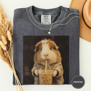 Puede incluir: Una camiseta de cuello redondo gris carbón con un estampado gráfico de un conejillo de indias marrón y blanco que lleva un suéter beige y sostiene un vaso de plástico transparente con una pajita. Etiqueta Comfort Colors en el cuello.