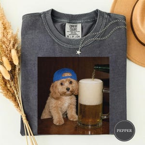 May include: A charcoal grey t-shirt with a Comfort Colors label at the neck. The graphic print features a light brown puppy wearing a blue baseball cap sitting next to a glass beer mug being filled with golden beer and a thick white foam head.