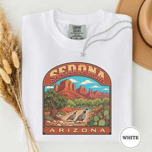 May include: A white t-shirt featuring a graphic print of the Sedona, Arizona landscape. The design includes red rock formations, a blue sky, a green tree, a cactus, and two quails on a path. The text reads Sedona Arizona in a retro font.