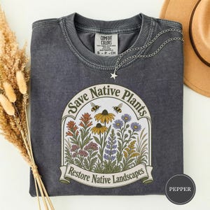 May include: A charcoal gray crewneck t-shirt featuring a graphic print of colorful wildflowers and two bees inside an arched frame. The text reads Save Native Plants and Restore Native Landscapes. A silver star charm necklace rests on the collar.