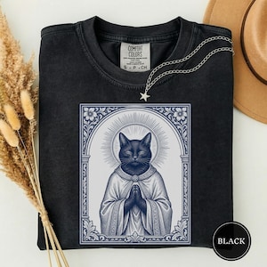 May include: A black crewneck t-shirt featuring a graphic print of a cat with closed eyes, wearing a robe with hands in a prayer position, framed by an ornate archway with a halo. The shirt has a Comfort Colors label at the inner neckline.