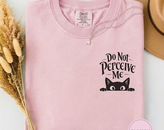 Peut inclure: Un t-shirt rose clair avec un motif noir représentant un chat qui dépasse au-dessus du texte "Do Not PERCEIVE ME" sur la poitrine. Le t-shirt a un col rond et une étiquette Comfort Colors à l'intérieur. Un collier étoile argenté repose dessus.