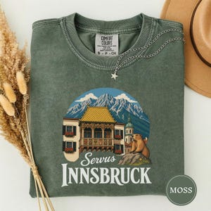 May include: A moss green crew neck t-shirt featuring a graphic print of the Golden Roof in Innsbruck, Austria, with mountains, a marmot, and the text "Servus INNSBRUCK". The shirt has a vintage wash finish and a silver star pendant necklace.