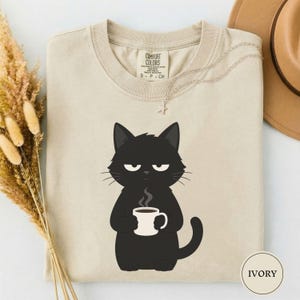 May include: A beige crew neck t-shirt featuring a graphic of a black cat holding a white coffee mug with steam rising. The shirt has a Comfort Colors brand label at the inner neckline and a small circular tag in the corner with the text IVORY.