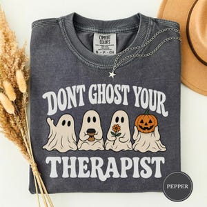 Puede incluir: Una camiseta gris carbón con lavado vintage que presenta el texto blanco DONT GHOST YOUR THERAPIST. El gráfico incluye cuatro fantasmas blancos: uno saludando, uno con una taza de café, uno con una flor y uno con una calabaza de Halloween.